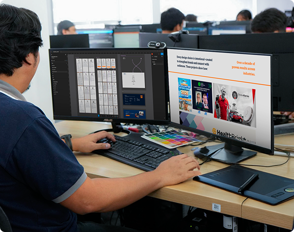 Graphic designer using a digital drawing tablet and ASUS dual-monitor setup to review a professional branding and marketing portfolio.